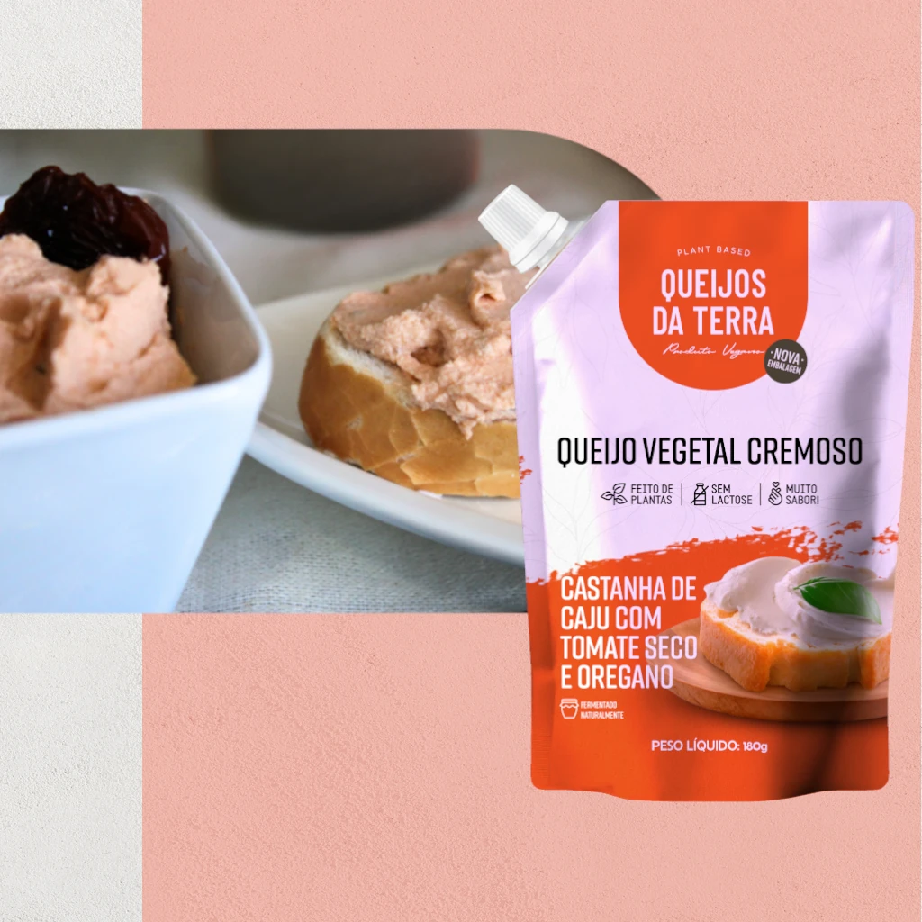 Queijo Cremoso de Castanha de Caju com Tomate Seco e Orégano. Em primeiro plano, está a embalagem de cor laranja vista de frente. Ao fundo, vê-se o queijo cremoso de cor laranja claro espalhado sobre um pedaço de pão e dentro de uma pequena tigela.