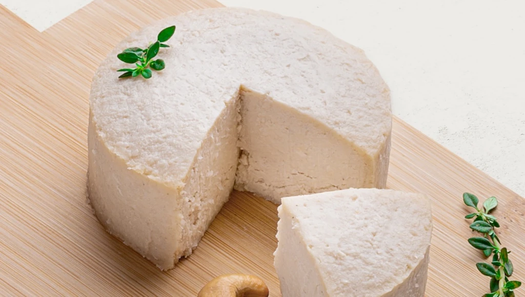 Queijo de Castanha de Caju com Gergelim em uma tábua de madeira, vista lateral. O queijo é lindamente branco.