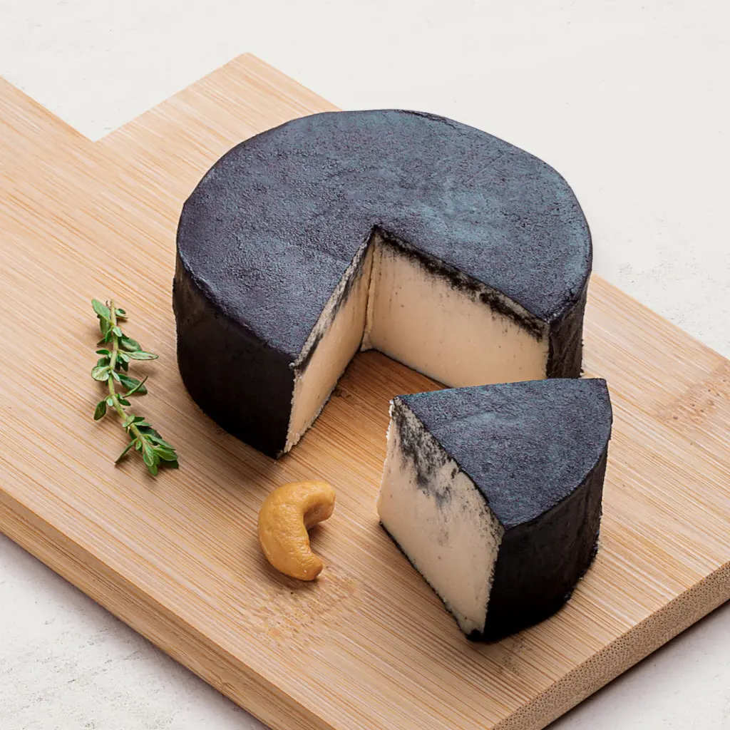 Queijo de Castanha de Caju Defumado em uma tábua de madeira, vista lateral. O queijo tem uma cor preta intensa.