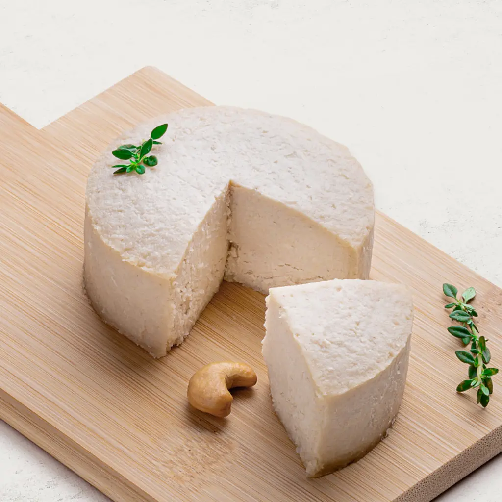 Queijo de Castanha de Caju com Gergelim em uma tábua de madeira, vista lateral. O queijo é lindamente branco.