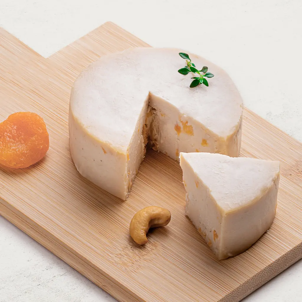 Queijo de Castanha de Caju Trufado com Damasco em uma tábua de madeira, vista lateral. Queijo de cor clara com pedaços de damascos visíveis.