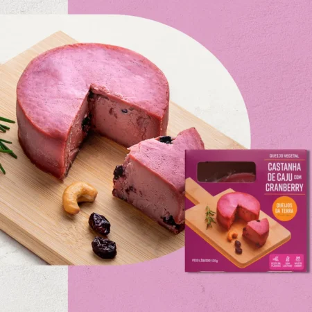 Queijo de Castanha de Caju com Cranberry sobre uma tábua de madeira, visto de lado. O queijo tem cor rosa. Em primeiro plano, sua embalagem roxa.