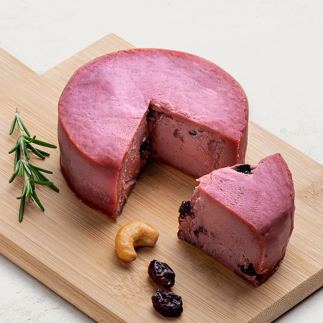Queijo de Castanha de Caju com Cranberry sobre uma tábua de madeira, visto de lado. O queijo tem cor rosa.