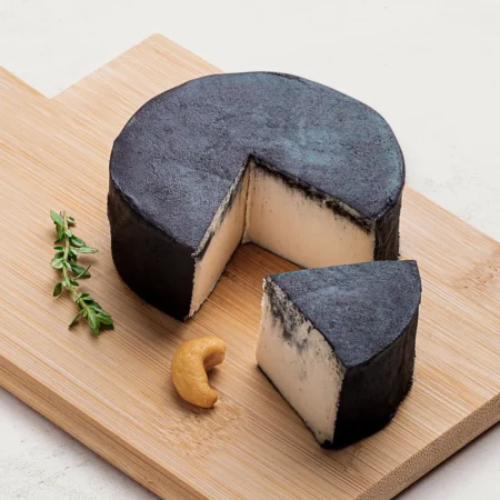 Queijo de Castanha de Caju Defumado sobre uma tábua de madeira, visto de lado. O queijo tem uma cor preta intensa.