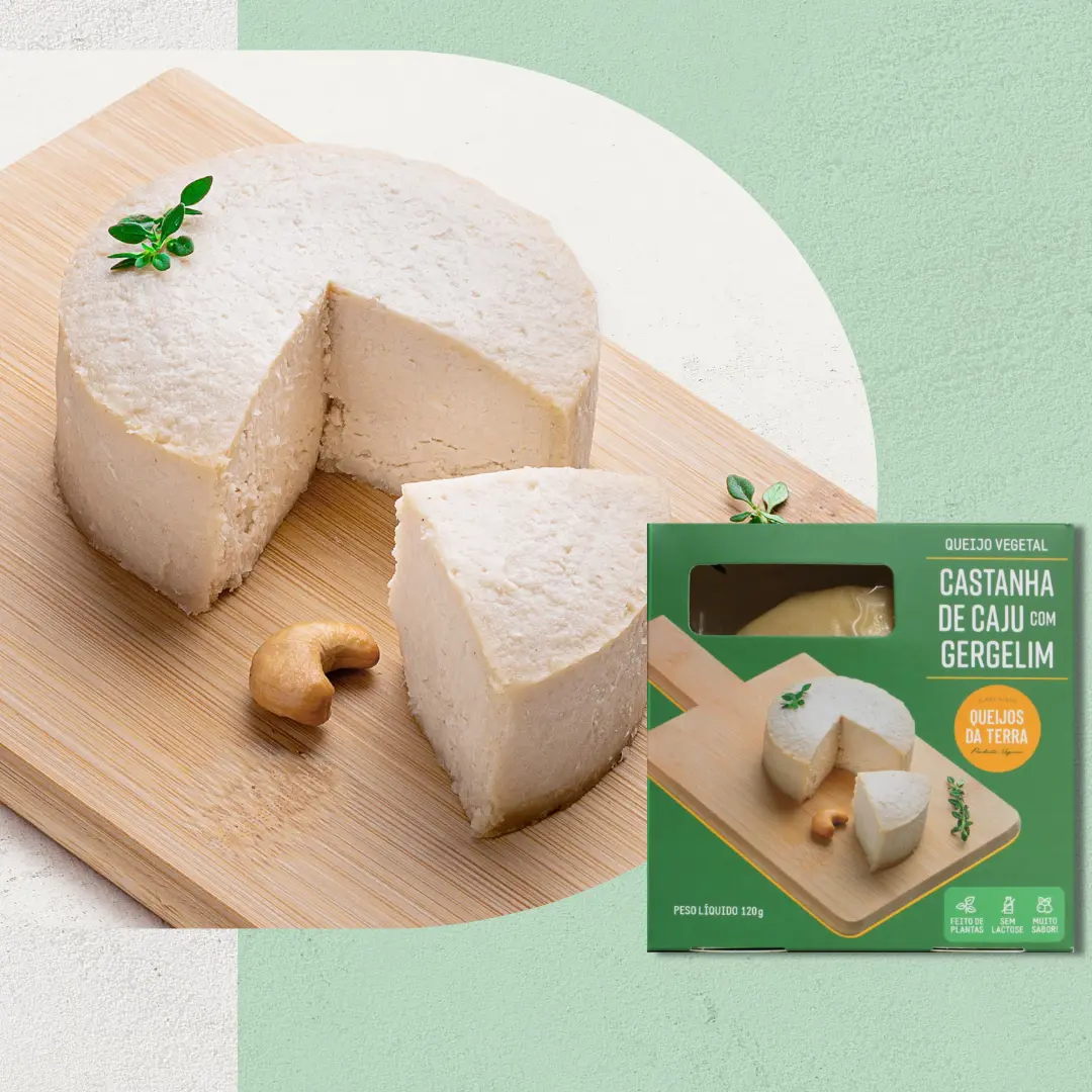 Queijo de Castanha de Caju com Gergelim sobre uma tábua de madeira, visto de lado. O queijo tem uma cor branca. Em primeiro plano, sua embalagem verde.