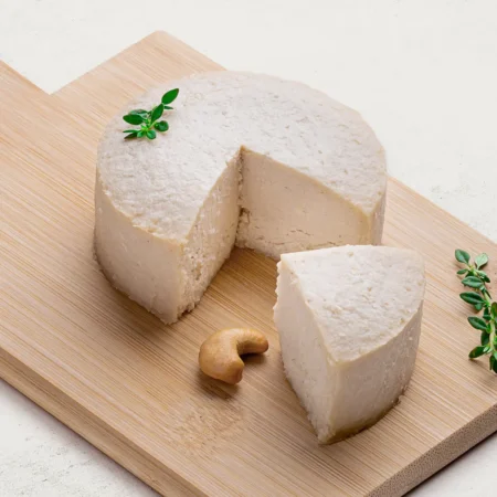 Queijo de Castanha de Caju com Gergelim sobre uma tábua de madeira, visto de lado. O queijo tem uma cor branca.