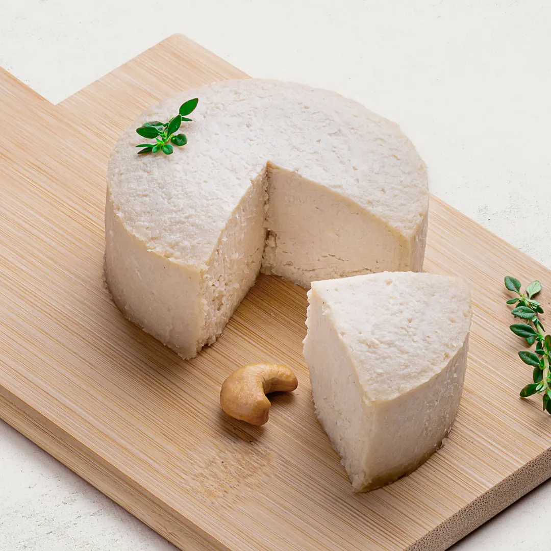 Queijo de Castanha de Caju com Gergelim sobre uma tábua de madeira, visto de lado. O queijo tem uma cor branca.
