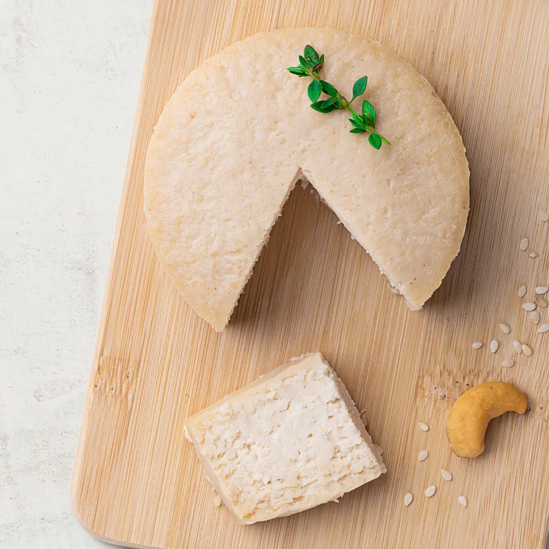 Queijo de Castanha de Caju com Gergelim sobre uma tábua de madeira, visto de cima. O queijo tem uma cor branca.