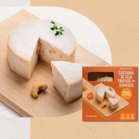 Queijo de Castanha de Caju Trufado com Damasco sobre uma tábua de madeira, visto de lado. O queijo tem uma cor clara com pedaços de damasco visíveis. Em primeiro plano, sua embalagem laranja.