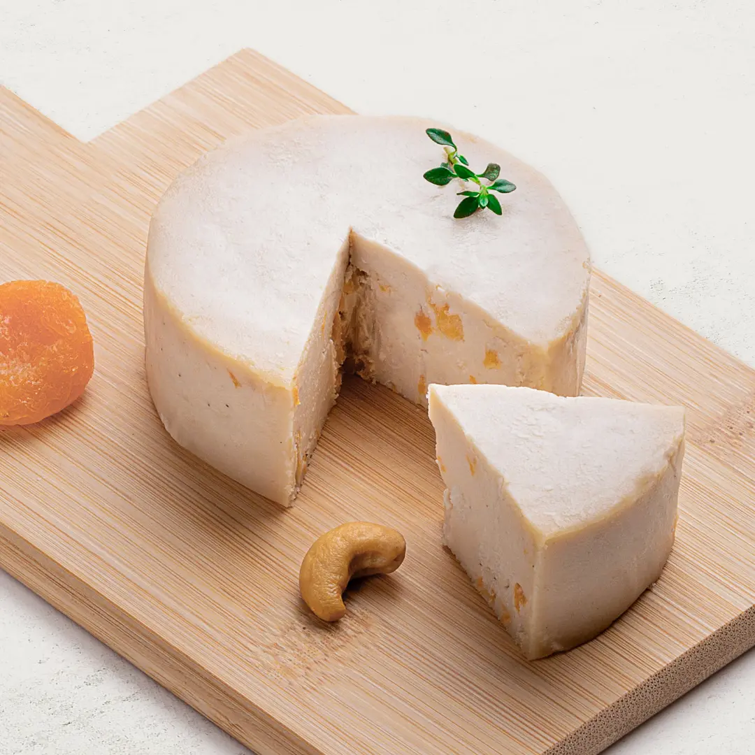 Queijo de Castanha de Caju Trufado com Damasco sobre uma tábua de madeira, visto de lado. O queijo tem uma cor clara com pedaços de damasco visíveis.